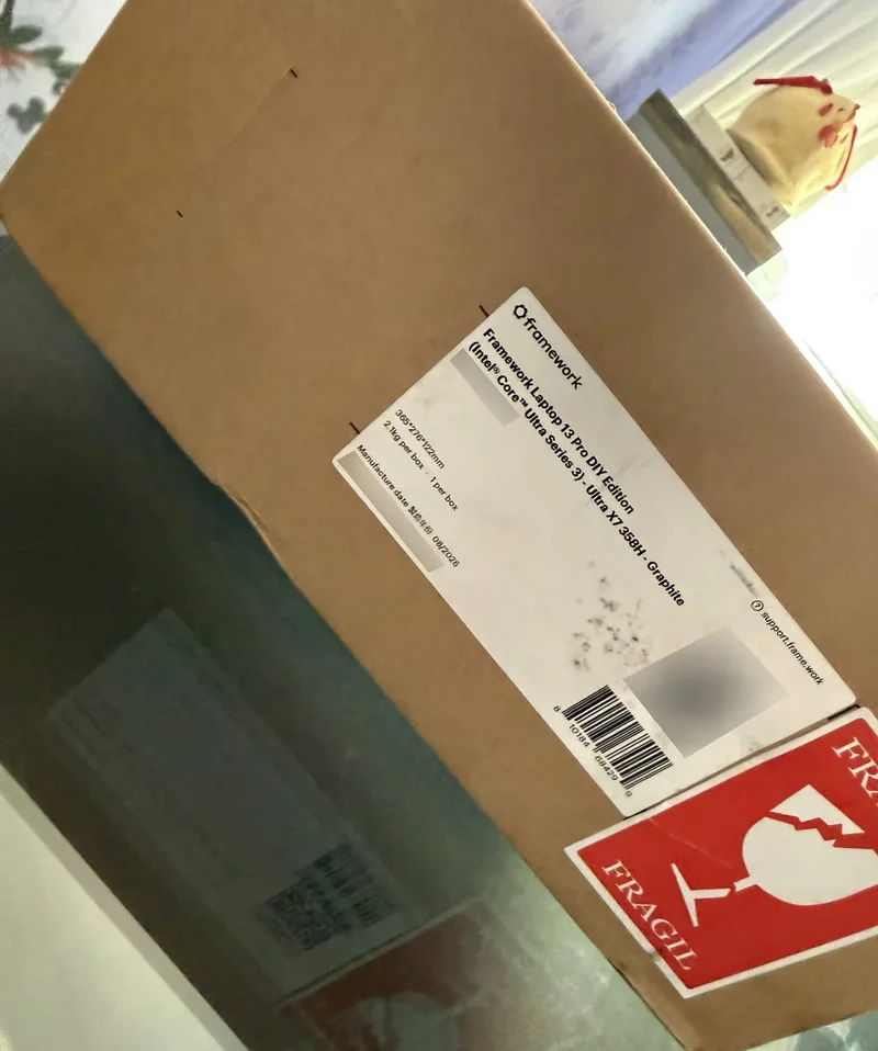 A cardboard box labeled as containing a Framework Laptop 13 DIY Edition (Intel Core Ultra Series 3) Ultra X7 235GH - Graphite with a visible 'Fragile' sticker.