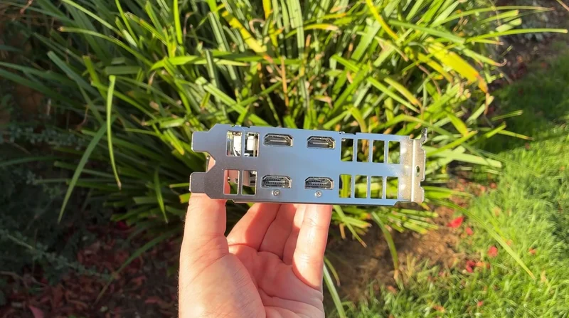 A person holds a GPU I/O bracket with three DisplayPort and one HDMI port against a background of greenery.