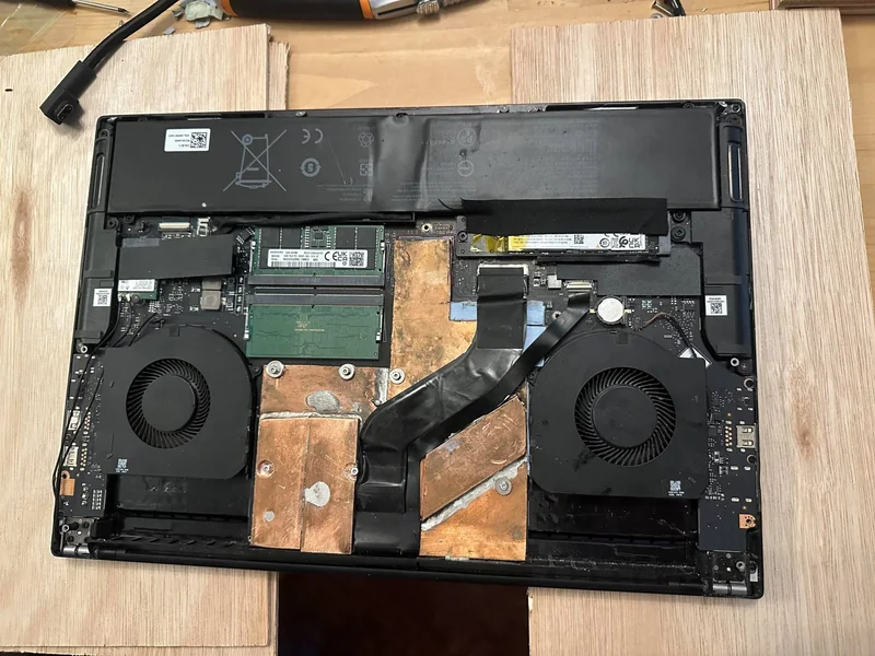 The underside of an open laptop showing dual cooling fans, a battery, RAM modules, and copper heat sinks.